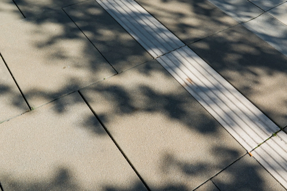 Nahaufnahme eines kalksteinfarbenen Betonsteins als Pflasterbelag, durchzogen von einem hellen Leitstreifen zur Orientierung, mit feiner Oberflächenstruktur und sichtbaren Licht‑Schatten‑Effekten