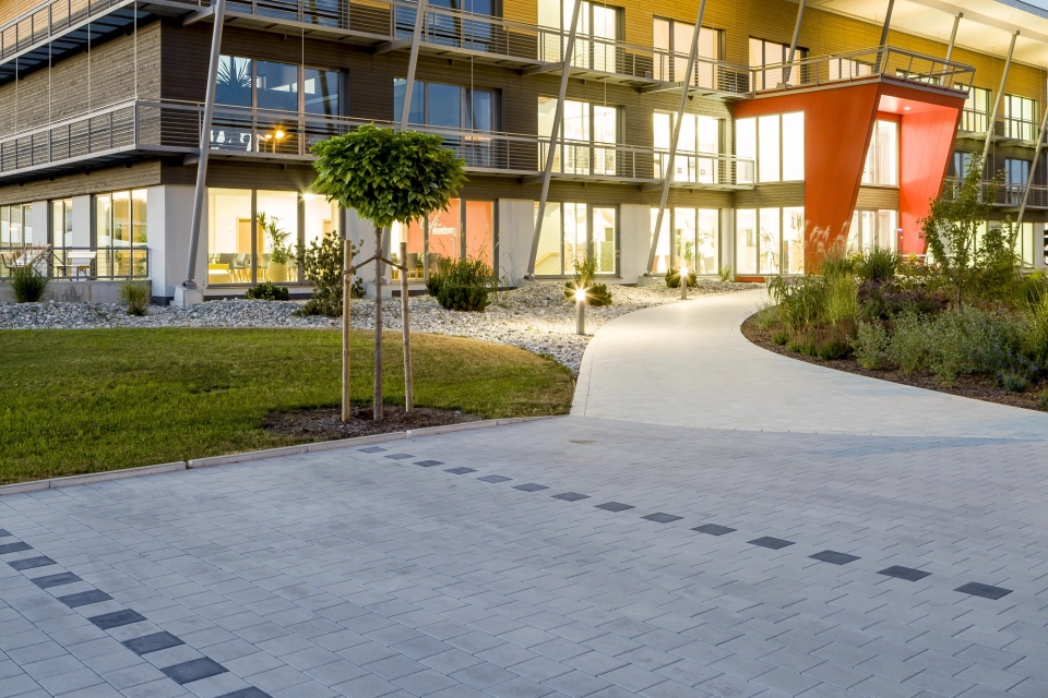 moderne Parkplatzgestaltung aus hellgrauem Pflaster mit geschwungenem Fußweg hin zu großem farbenfrohen Unternehmensgebäude