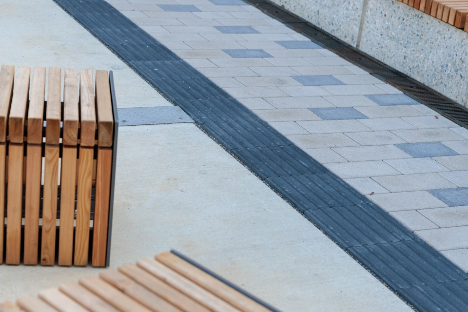 Exterior area with light beige paving, into which occasional grey paving elements are incorporated as accents. A dark tactile strip runs along the side. In the foreground is seating made of wooden slats with metal frames, behind it a long bench in front of a wall of vertical wooden slats.