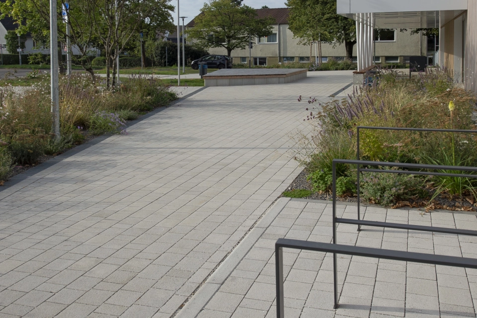 Path made of grey-lime, permeable climatic stones. Next to it are anthracite-coloured bicycle stands, shrubs and street lamps.
