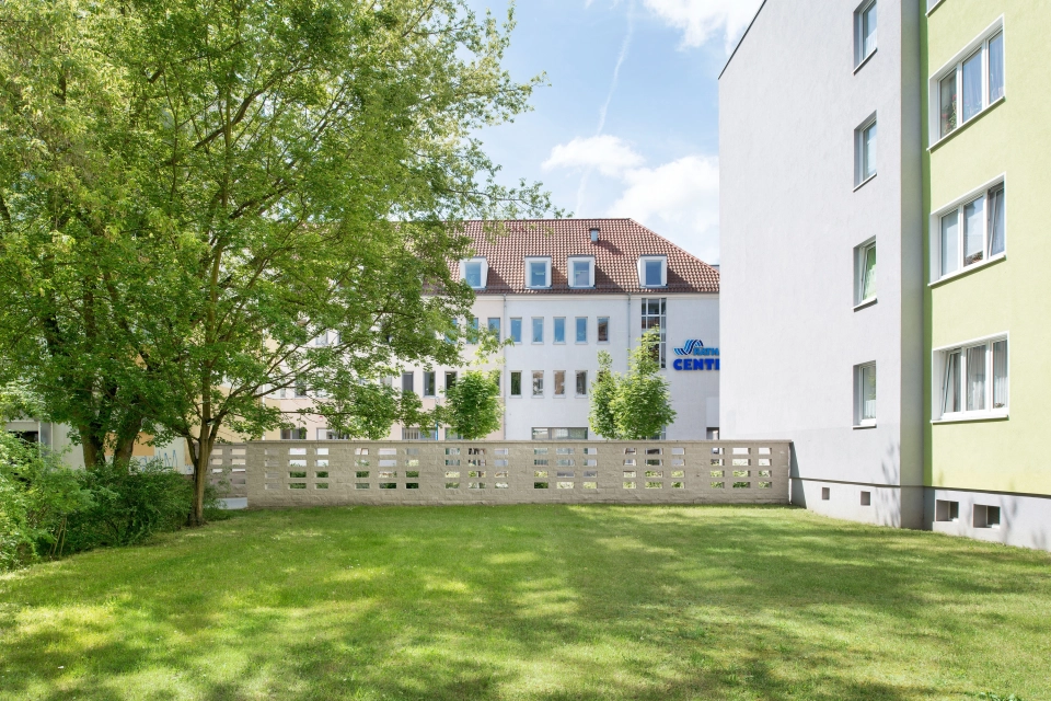 lange sandfarbene Mauer auf Rasenanlage neben vierstöckigem weiß-gelben Gebäude und Bäumen