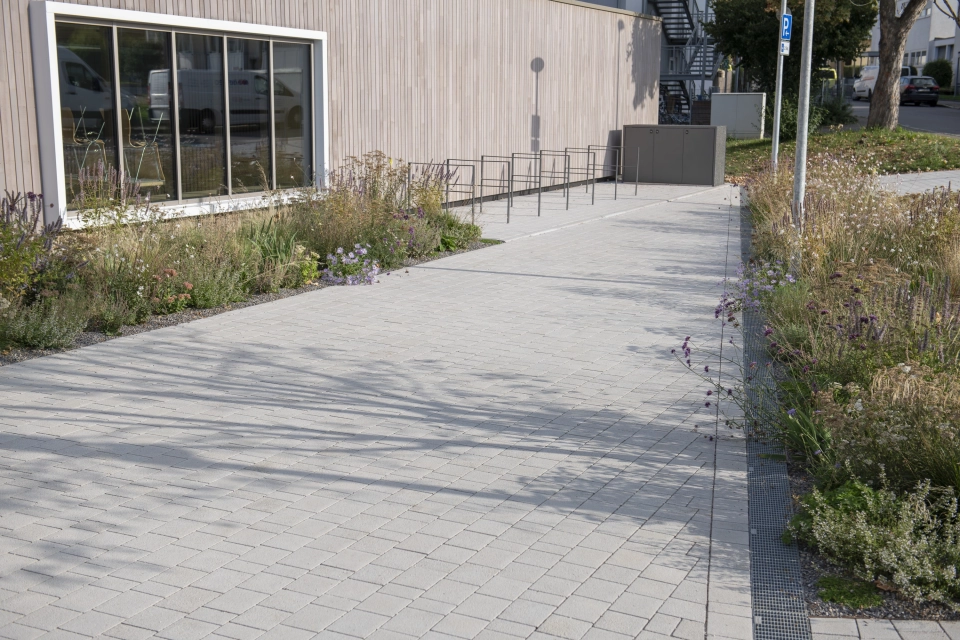 Path made of lime-grey, permeable climate stones. Next to it are shrubs and grasses and in the background are anthracite-coloured bicycle stands.