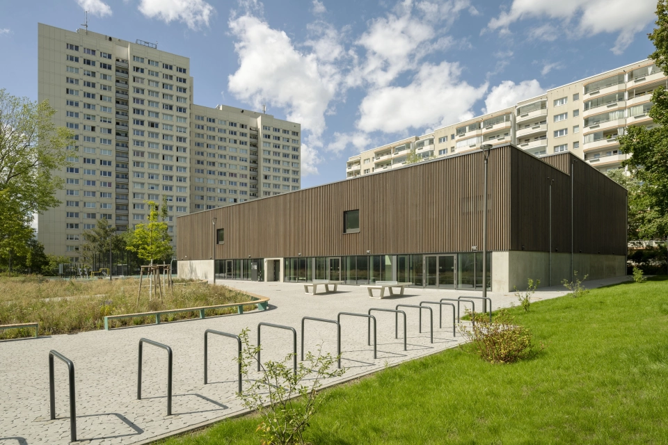Ein großes Schulgebäudes mit Holzfassade mitten in einer Stadt. Vor dem Gebäude liegt ein Schulhof mit vielen Fahrradständern, Tischtennisplatten und begrünten Flächen.