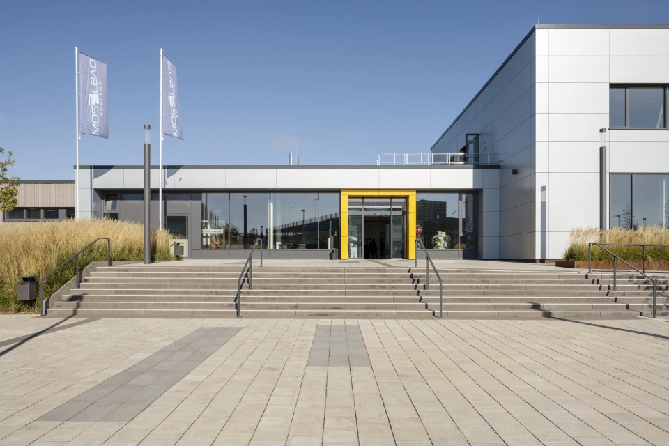 Großer Vorplatz mit grauem GDM.SCENIC individual und dunkleren Betonsteinen zur gestalterischen Auflockerung. Eine Treppe führt auf das große Gebäude mit grauer Fassade zu. Um den Eingangsbereich herum ist eine auffällige gelbe Umrandung.