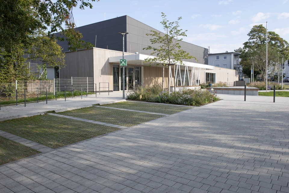 Modern exterior façade of a sports hall with anthracite-coloured panels in the upper area and wooden cladding in the lower area. In front of it is a forecourt paved with grey-lime, permeable concrete blocks with parking spaces made of grass pavers and shrubs, grasses and trees. 