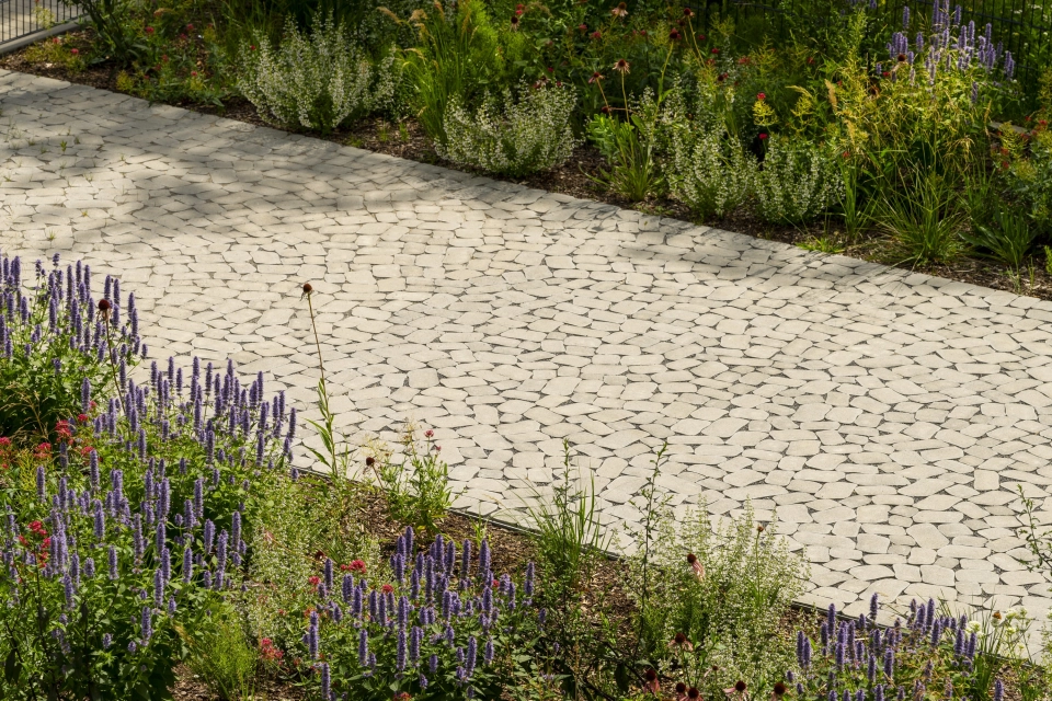 Ein mit natürlichen wirkenden, rundlichen Betonsteinen gepflasterter Weg, welcher mitten durch wild und bunt bewachsene Blumenbeete führt.