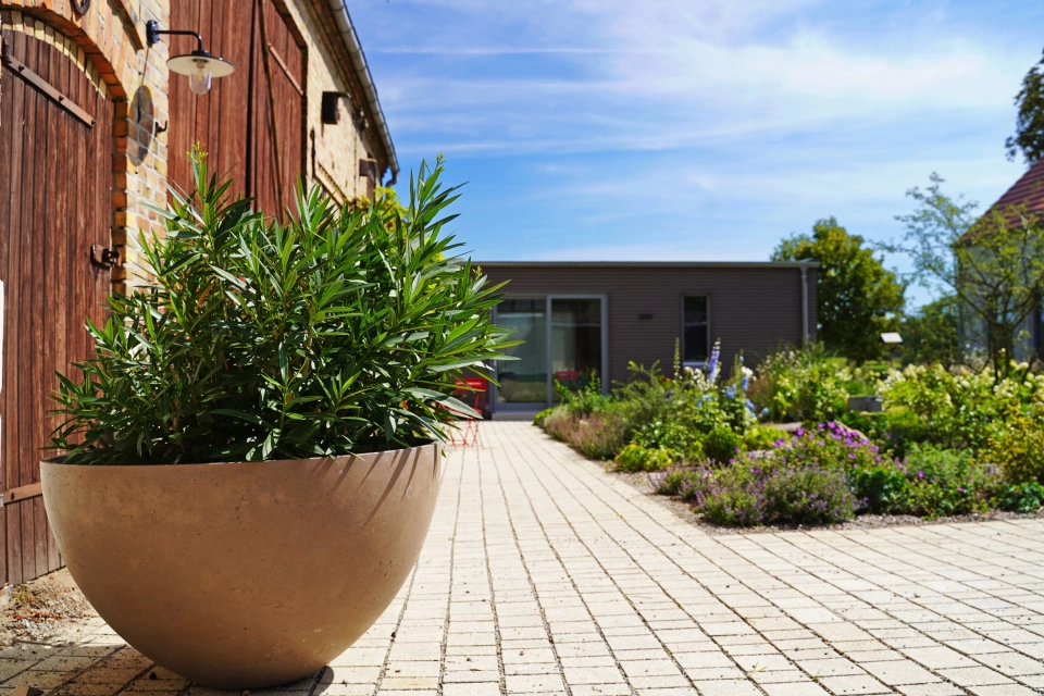 On the left of the picture is a rustic brick building with large wooden gates and a large planter in the foreground. On the right is a neatly landscaped garden planted with flowers and shrubs. In front of the garden is a natural beige paved courtyard laid out in rows. A modern building with large glass doors can be seen in the background.