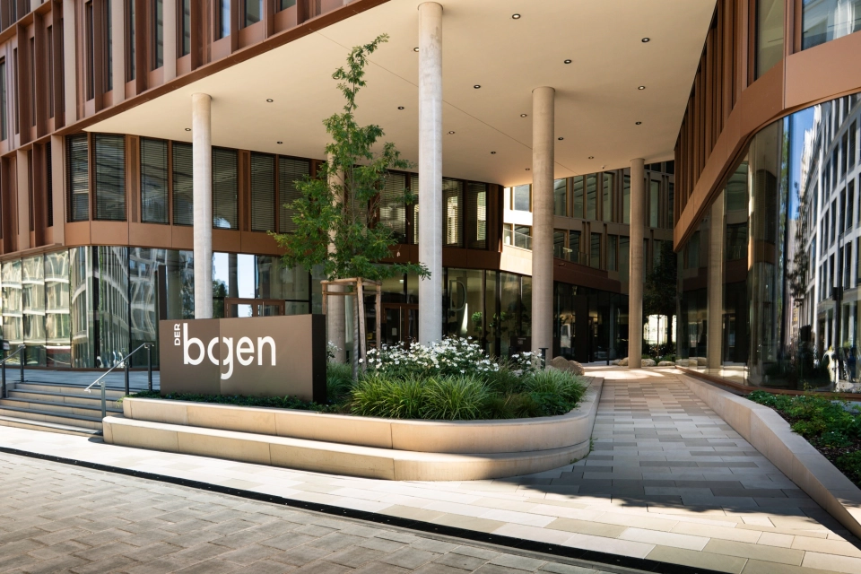 Blick auf den Eingangsbereich des Gebäudeinnenhofes. Hier ist die Fläche mit GDM.SCENIC individual in verschiedenen Kalksteinfarbtönen gepflastert. Das Gebäude rundherum hat viele große Fenster und wird zusätzlich durch mehrere hohe Säulen gehalten. Außerdem ist ein großes Beet mit verschiedenen Sträuchern und Bäumen und ein schwarzes Schild davor zu finden.