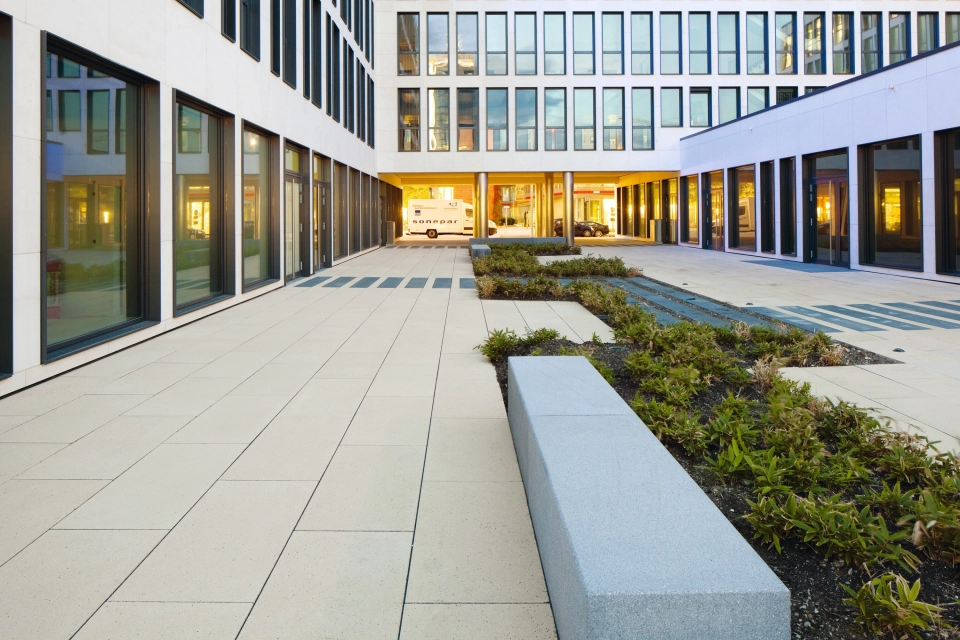Begrünter Innenhof eines modernen Geböudekomplexes mit Sitzelementen und großflächigem Plattenbelag