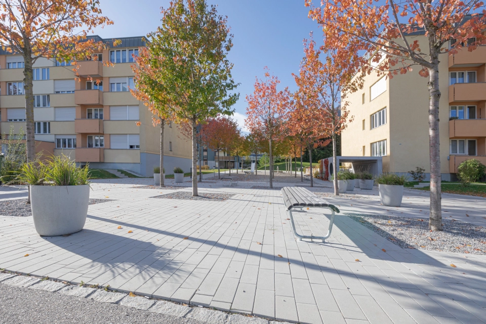 breiter hellgrau gepflasterter Platz mit Bäumen, Sitzbänken und Büschen vor zwei orangenen Mehrfamilienhäusern 