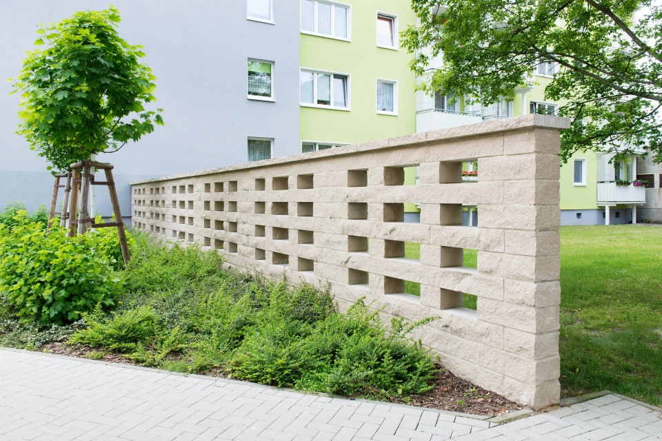 hohe sandfarbene Mauer mit fensterähnlichen Aussparungen hinter Fußgängerweg und bepflanztem Grünstreifen