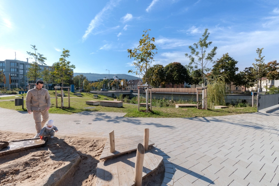 Openly designed outdoor area with a path made of concrete blocks bordering a sand play area with wooden elements. In the foreground is an adult with a small child, in the background are green areas, young trees and a watercourse with plants on the bank.