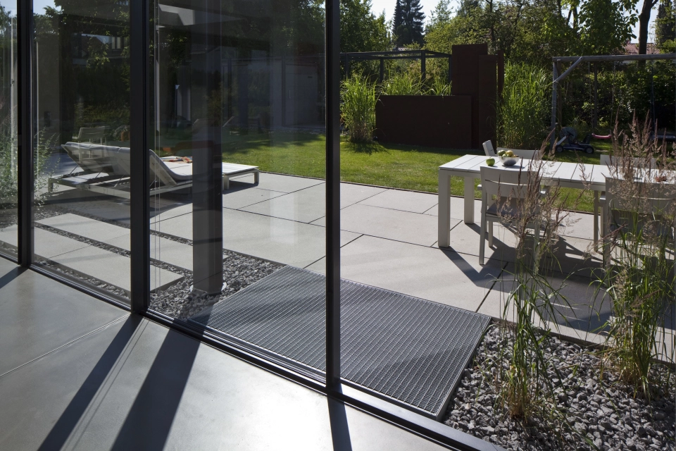 Glasfront eines Wohnhauses mit Blick auf den Garten und die Terrasse mit Esstisch und breitem Liegestuhl