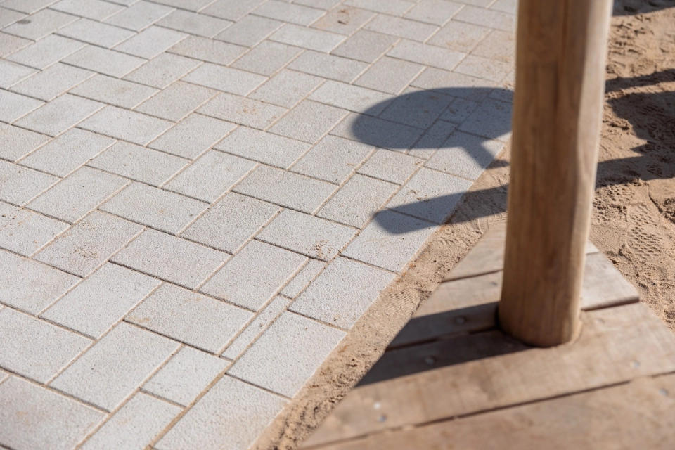 Close-up of a light, beige-coloured paving made of GDM.KLIMASTEIN. On the right of the picture are a wooden post and a wooden edging, next to it is a neighbouring sandy ground area.