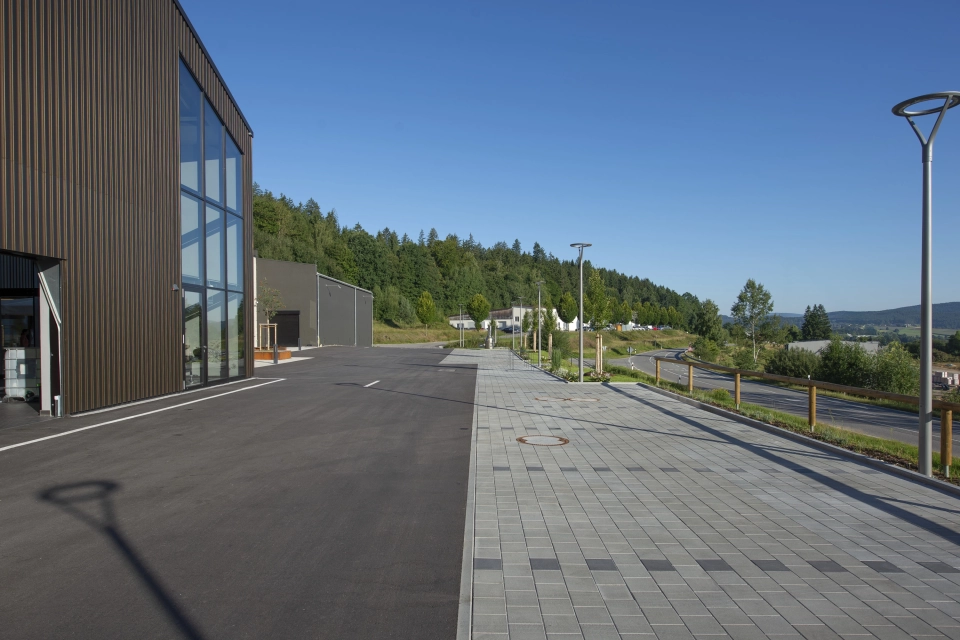 Rechts befindet sich eine Parkfläche aus grauem, versickerungsfähigem Betonstein, neben einer geteerten Straße und eine Holzfassade mit großer Fensterfront. Die einzelnen Parkplätze sind durch dunkelgraue Betonsteine abgetrennt.