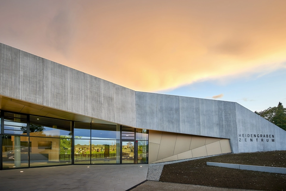 Moderne Gebäudefassade in Holzoptik mit großer Fensterfront und beigen Designelementen. Der Platz davor ist mit fünfeckigen, kalksteinfarbenen Betonsteinen gepflastert. Daneben ist eine Fläche aus Erde.