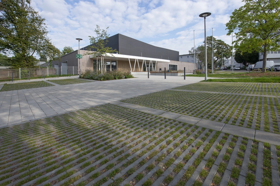In the foreground is a car park made of green grass paving stones. Behind it is a path made of concrete blocks and a modern sports hall with an anthracite-coloured façade and wooden cladding. Surrounding it are green trees and shrubs.