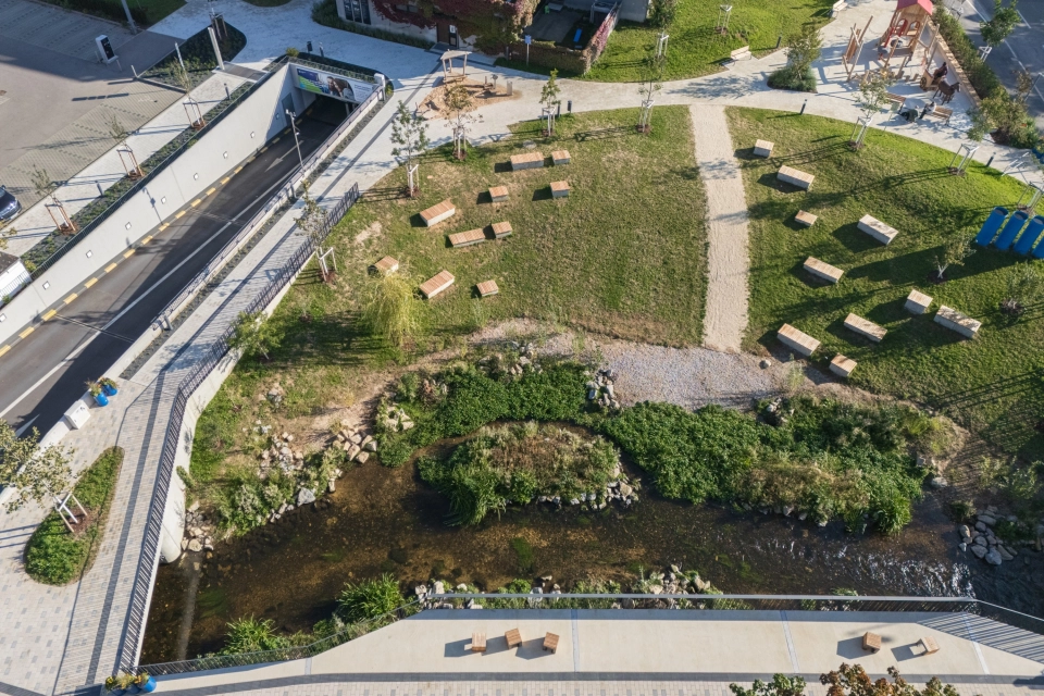 Aerial view of a newly designed park area with paved paths, a small stream, green areas with many benches and neighbouring play and traffic areas.