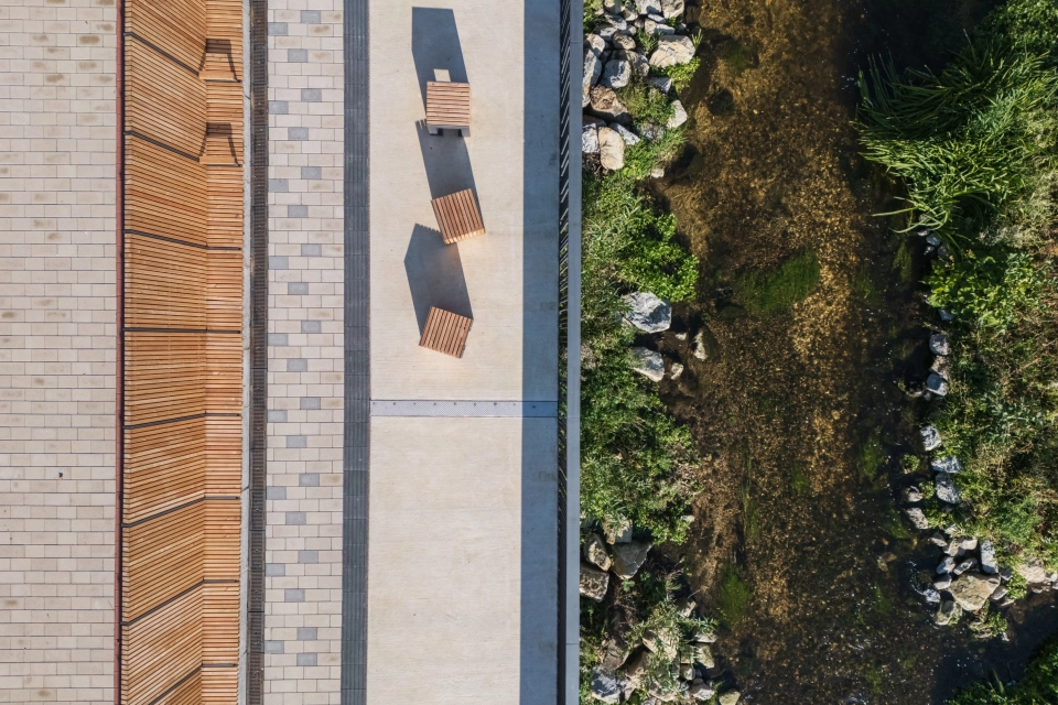 Top view of a riverside path with several wooden benches, adjacent to a natural stream with stones and dense bank vegetation. The path is paved with beige GDM.KLIMASTEIN.