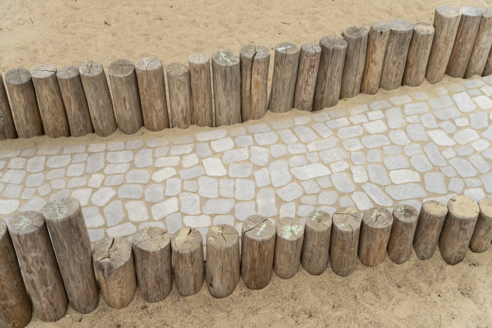 Mit GDM.BOLERO gepflasterter Weg, eingefasst von Rundholzstämmen. Naturnahe Wegeführung zwischen Spiel- und Aufenthaltsbereichen.