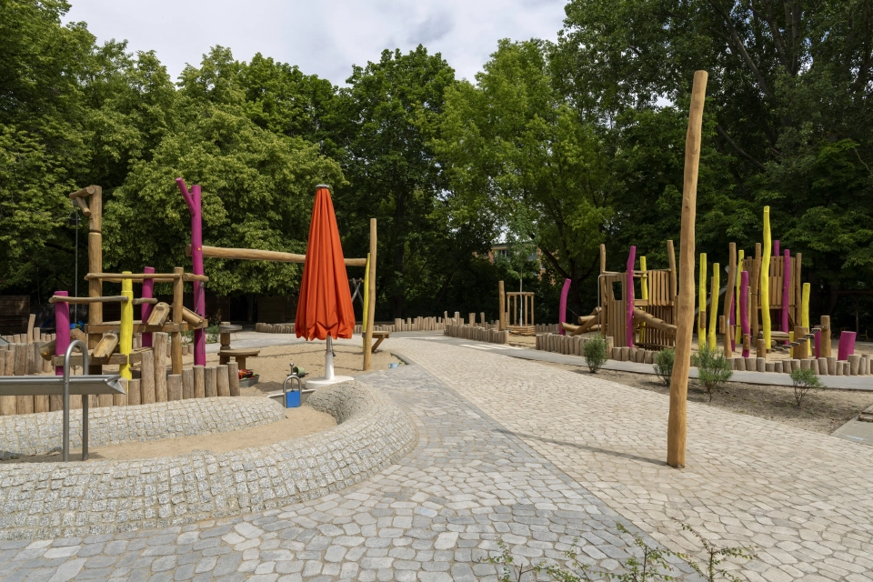 Außenanlage der Kita Wolkenschaf mit zweifarbiger GDM.BOLERO-Pflasterung, Naturholz-Spielgeräten, großem Baumstamm und orangefarbenem Sonnenschirm.