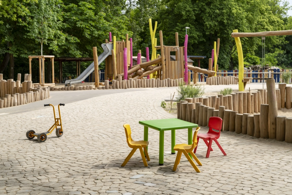 Grüner Kindertisch mit zwei gelben und einem roten Stuhl auf GDM.BOLERO. Links ein gelber Kinderroller, dahinter Spielgeräte aus Naturholz.