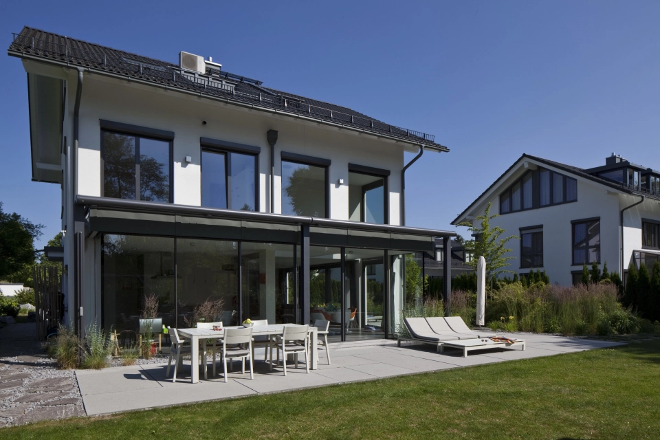 Modernes Einfamilienhaus mit Wintergarten und Glasfront vor heller Terrassenfläche mit Esstisch und Liegestuhl