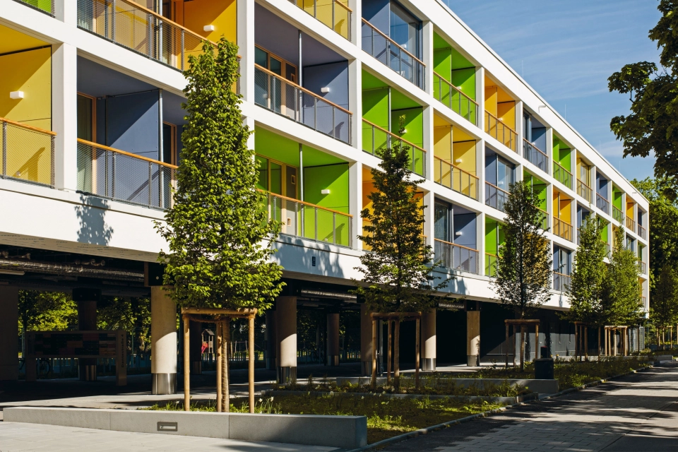 Wohngebäude mit blauen, grünen und gelben Balkonen vor Parkanlage mit Bäumen